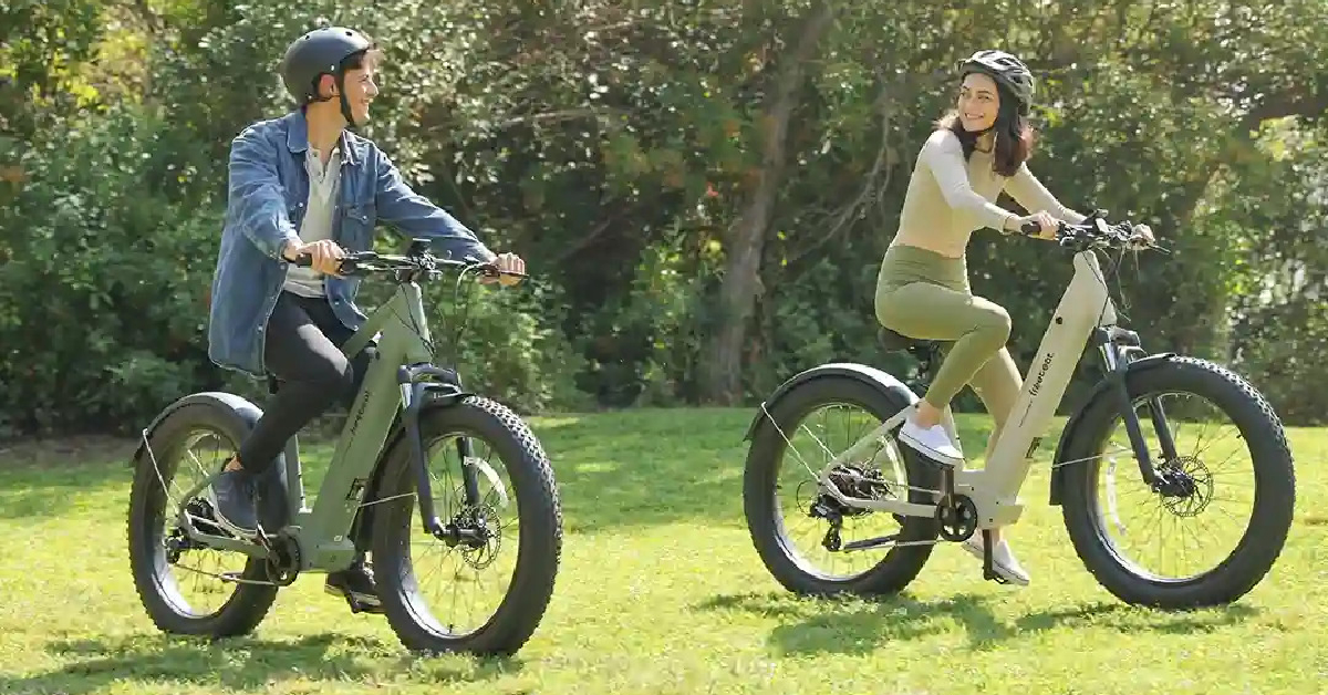 Two people are riding Mountain E-Bikes on a grassy field. Both are wearing helmets, and the background hints at future trail adventures in a wooded area.