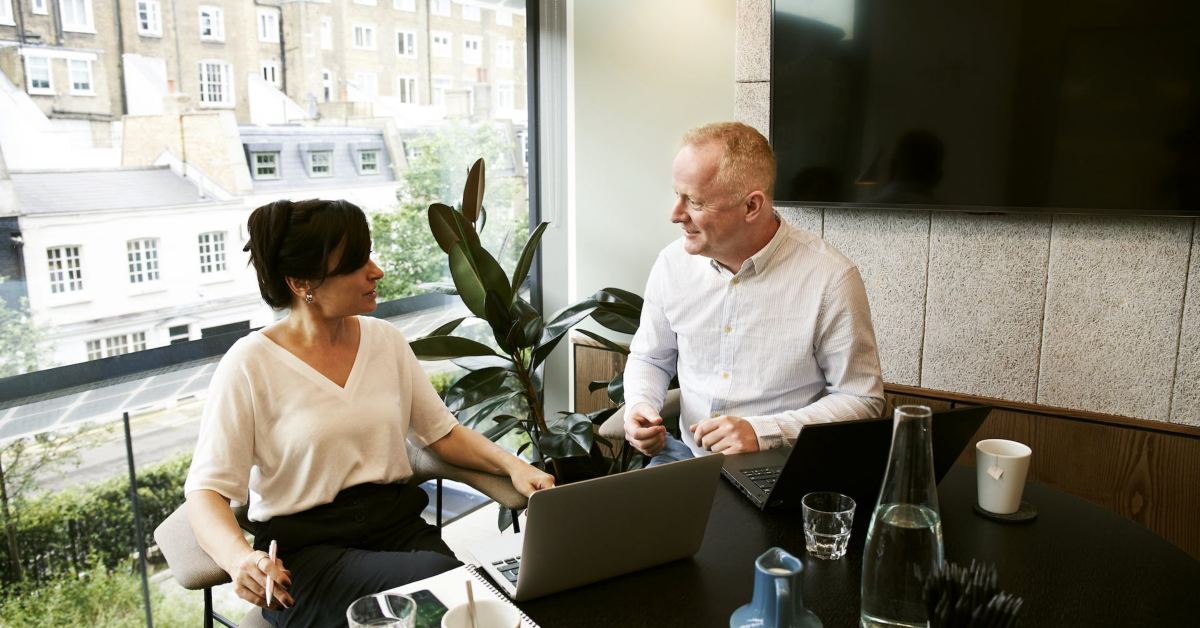 Two people sitting at a table with laptops in front of a window, collaborating on eProcurement for enhanced savings and efficiency.