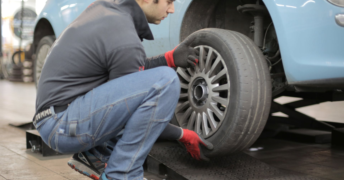 A mechanic in a gray sweatshirt and jeans, wearing red gloves, is installing a tire on a car lifted on a hydraulic jack, demonstrating essential car care tips for improving vehicle longevity.