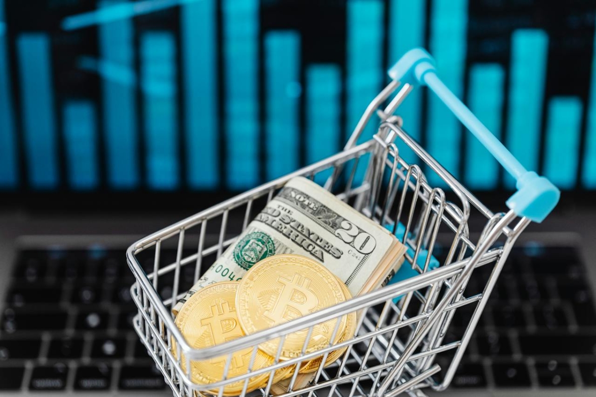 A miniature shopping cart containing US dollar bills and physical Bitcoin tokens sits on a laptop keyboard displaying Frontnode’s financial charts in the background.