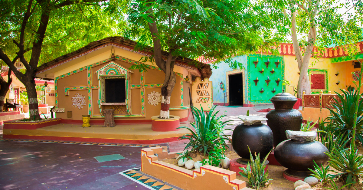 A vibrant courtyard within an Indian villa, adorned with traditional painted houses, large black pots, green plants, and shading trees. The scene is brought to life with colorful details and various geometric patterns gracing the buildings.