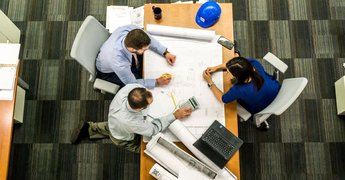 Three people at a table review large blueprints; one with a yellow highlighter, another with a calculator, and the third with a blue hard hat—essential techniques to strengthen business operations.