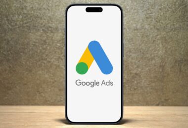 A smartphone showcasing the Google Ads logo stands upright on a wooden surface, set against a textured gray background. Perfect for e-commerce sales, this tool empowers businesses to create targeted campaigns that enhance visibility and drive customer engagement.