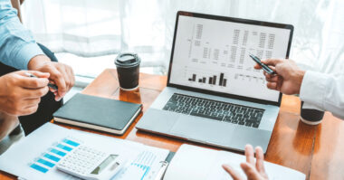 Three people discuss financial data displayed on a laptop screen at a table. Documents, a notebook, pen, calculator, and a coffee cup are present. Their insights could be a game-changer in this meeting.