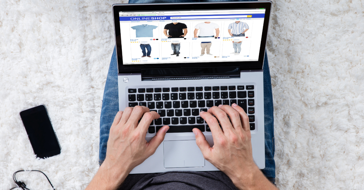 A person is browsing an online clothing store on a laptop, scrolling through an array of shirts and jeans. The laptop rests on their lap while a smartphone, possibly for checking tips for making money fast on eBay, is nearby on the carpeted floor.