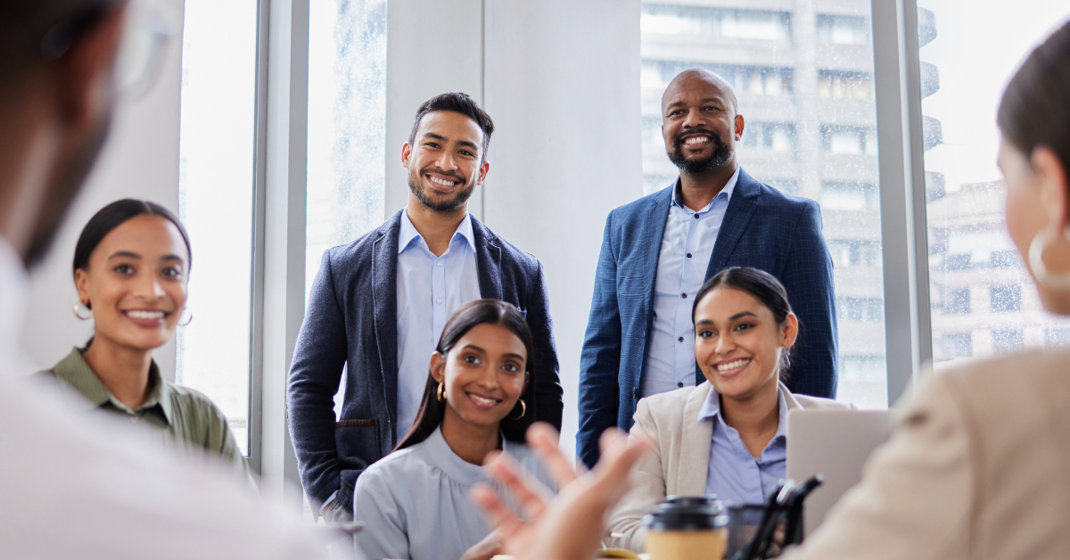 A group of six people in business attire are smiling and discussing effective leadership in an office setting with large windows in the background.