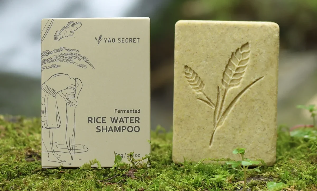 A box labeled "Fermented Rice Water Shampoo" rests beside a Rice Water Bar Shampoo atop the moss-covered ground, suggesting a natural way to store and maintain hair health.