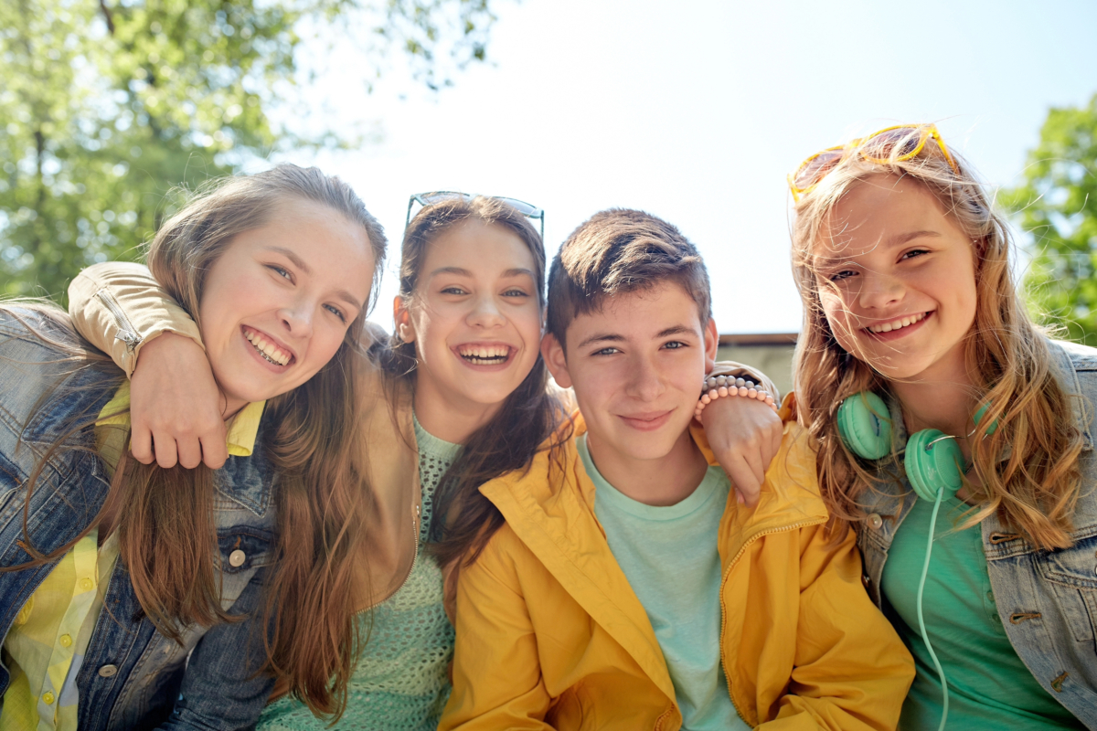 Four smiling teenagers, two girls on the left and two on the right, pose together outdoors. One girl has headphones around her neck, perhaps taking a break from her latest teen side hustle. They're all wearing casual, brightly colored clothing that reflects their vibrant energy.