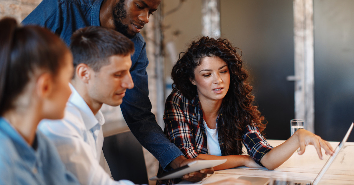 Four people collaborate at a table, discussing ecommerce trends on a laptop. One person holds a tablet, browsing their Shopify store for insights.