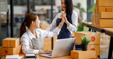 Two women high-five near a laptop, surrounded by stacked cardboard boxes in an office setting, celebrating their practical approaches to navigating the competitive ecommerce market.
