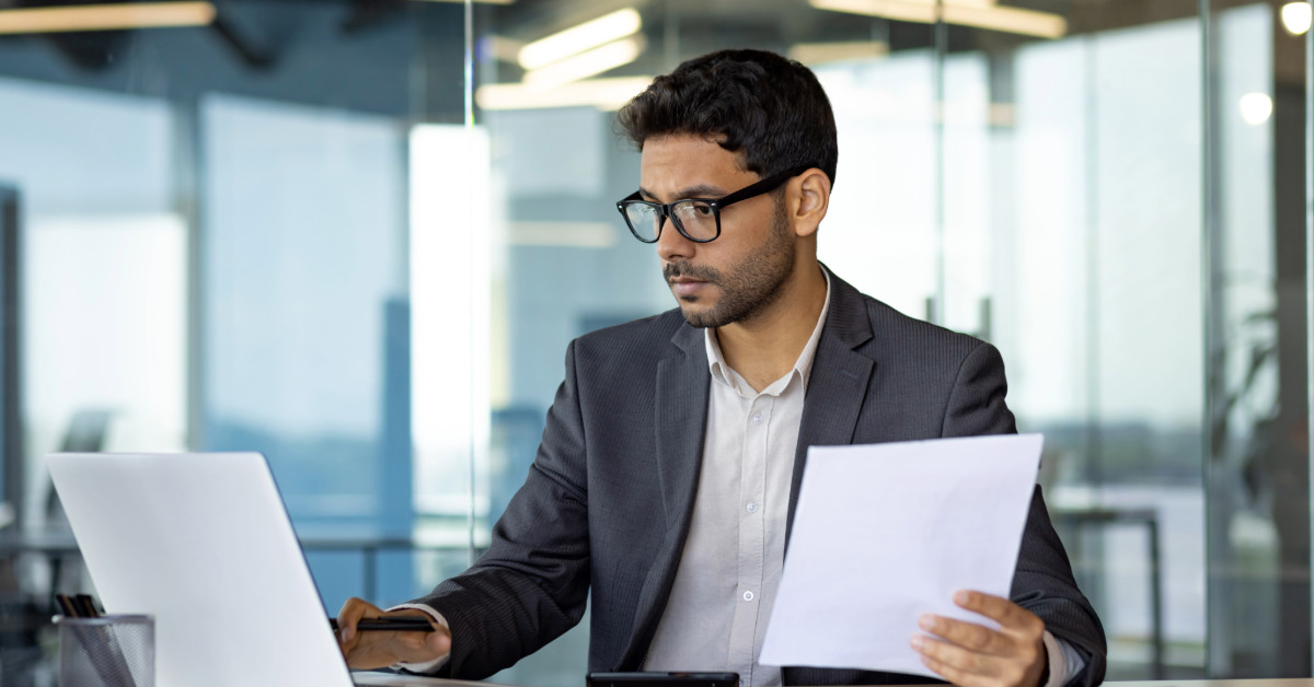 A man in a suit is sitting at a desk, analyzing risk management strategies on his laptop while holding a document. He is in a modern office with large windows, possibly working for one of the UAE investment companies.