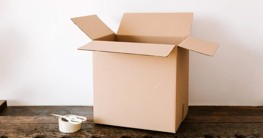 An open cardboard box sits on a wooden surface with a roll of tape and a pair of scissors beside it, an essential guide for global shippers.
