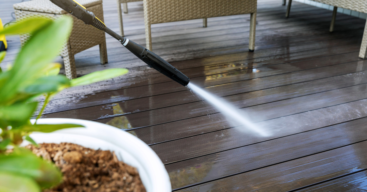 A person is using a pressure washer to clean a wooden deck beside patio furniture and a potted plant, ensuring every inch of the surface gleams. The efficient cleaning restores the inviting look, though opting for composite decking could reduce maintenance efforts in the future.