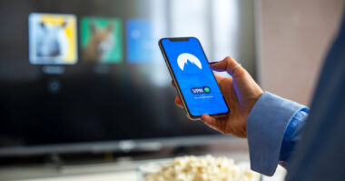 Person holds a smartphone displaying a VPN app interface, possibly dealing with common VPN problems, with a television screen showing various icons in the background and a bowl of popcorn in the foreground.