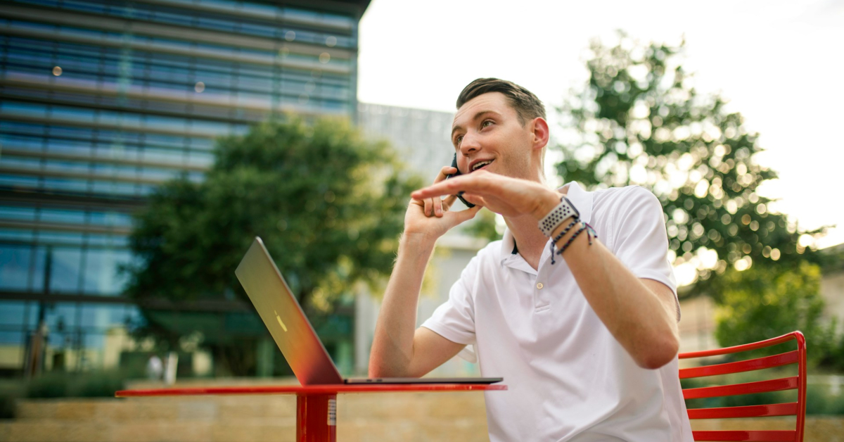 A person in a white shirt talks on a cellphone, likely providing tailored answers for customer support, while waiting at a red table with an open laptop outdoors. Trees and a modern building create a serene background.
