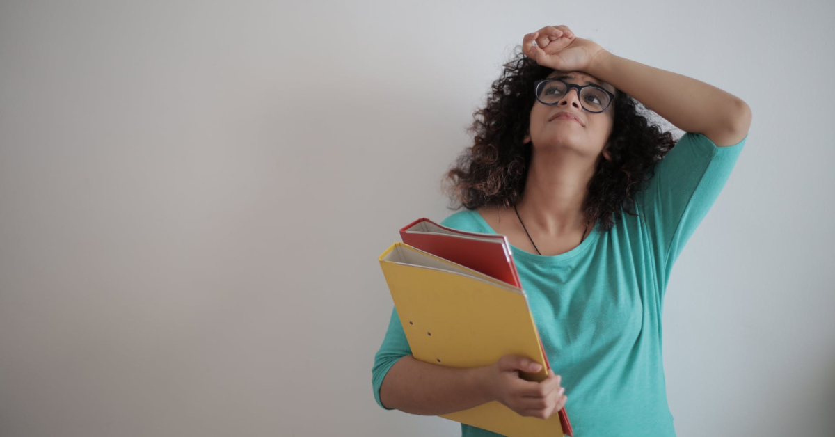 A person in a teal shirt is leaning against a white wall, holding yellow and red folders. Looking up with one hand on their forehead, they seem deep in thought about career challenges.