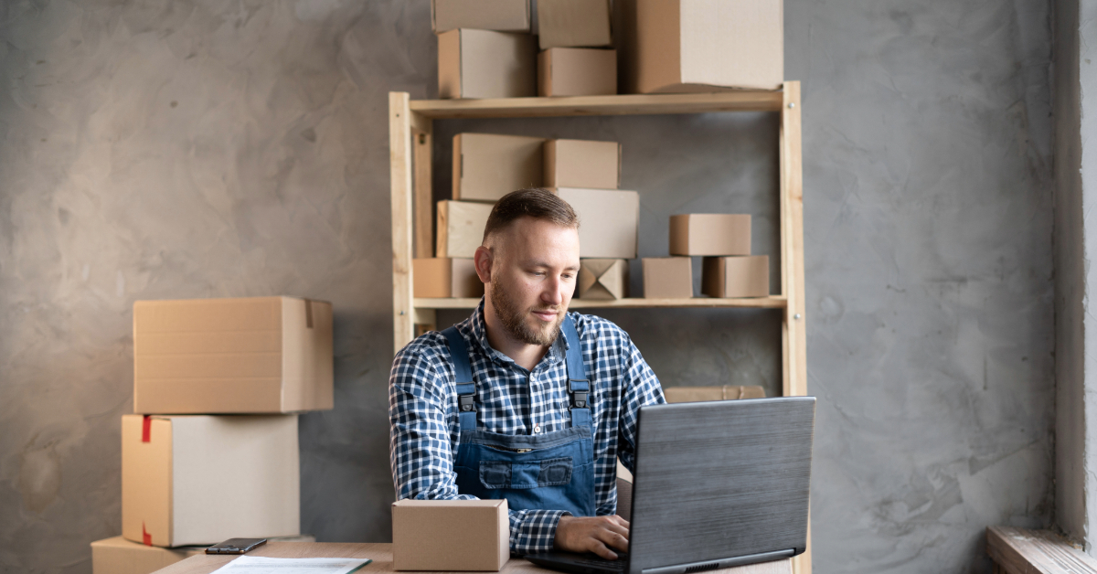 A man in a plaid shirt and overalls diligently works on his laptop at a wooden table, setting up his Shopify store. Cardboard boxes, filled with products for his DTC ecommerce business, are neatly stacked on shelves and the table against a gray wall.