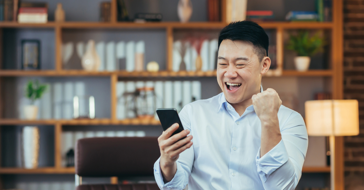 A person in a white shirt happily looks at their smartphone, raising a fist in excitement, as if they've just cracked the mystery of color prediction games, in a room with a bookshelf and lamp.