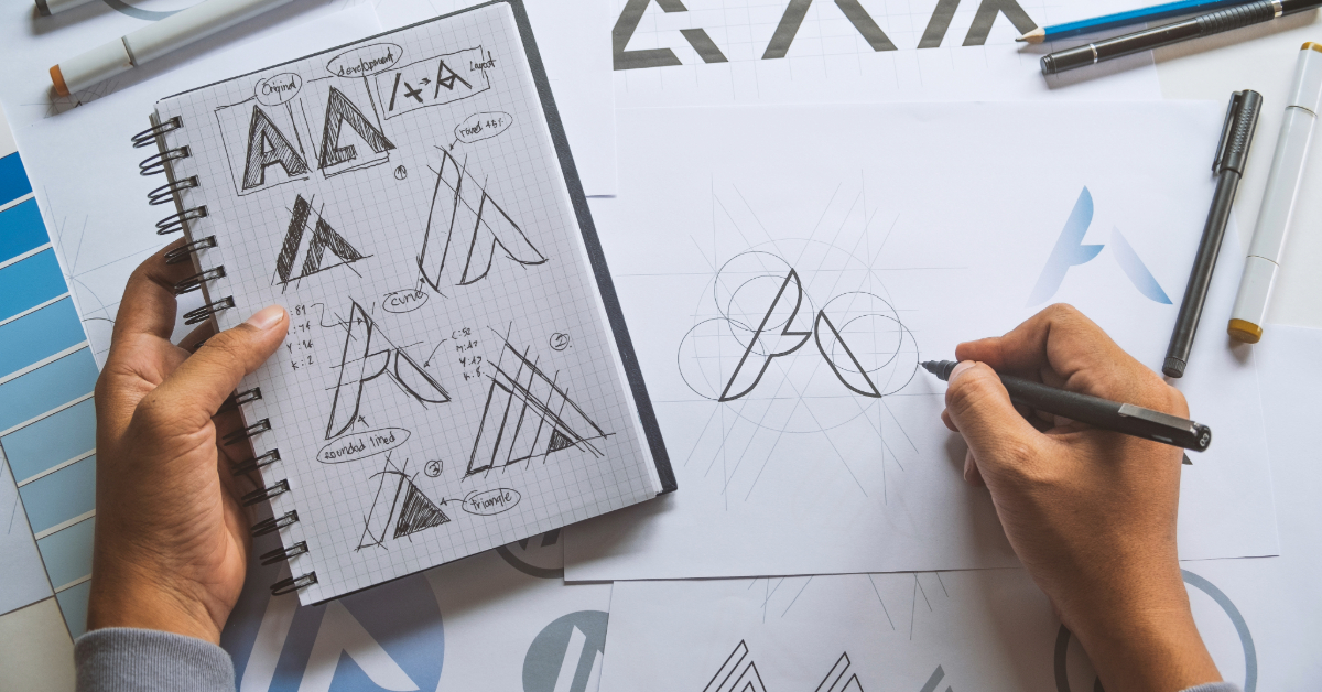 Designer sketching logo concepts on paper and in a notebook, surrounded by drafting tools, capturing the essence of DTC branding for an emerging Shopify ecommerce venture.