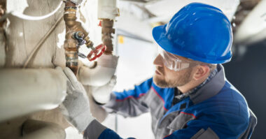 In an industrial setting, a worker wearing a blue hard hat and safety goggles examines a pipe amid surrounding plumbing, ensuring everything flows as smoothly as a Shopify-powered DTC ecommerce operation.