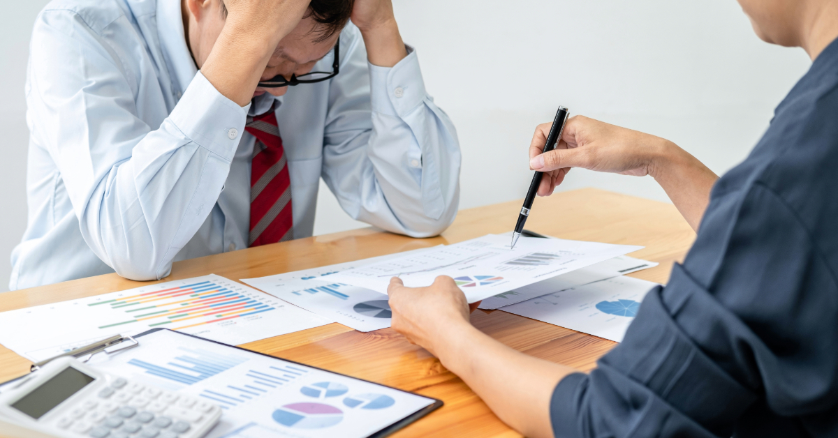 Two people at a table analyze ecommerce financial documents with charts. One looks stressed, while the other points at a document with a pen. A calculator and Shopify reports are visible on the table, hinting at the challenges of managing a DTC business model.