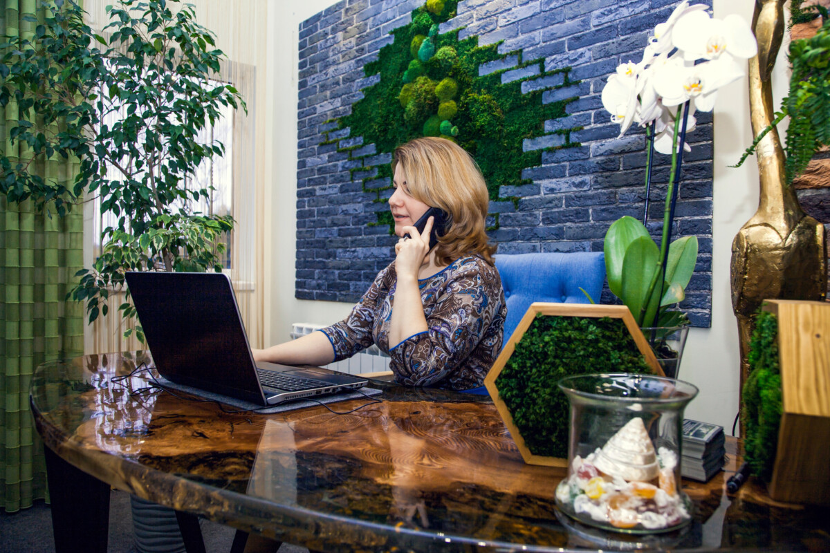 A woman sits at a wooden desk, chatting on the phone with an open laptop displaying her Shopify dashboard. The backdrop highlights a brick wall adorned with green moss artwork and lush plants, creating an inspiring environment for her thriving ecommerce venture.
