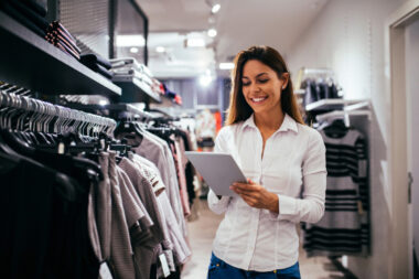 A woman in a white shirt uses a tablet while standing among clothing racks in a retail store, seamlessly executing an innovative strategy for efficient store fulfillment.