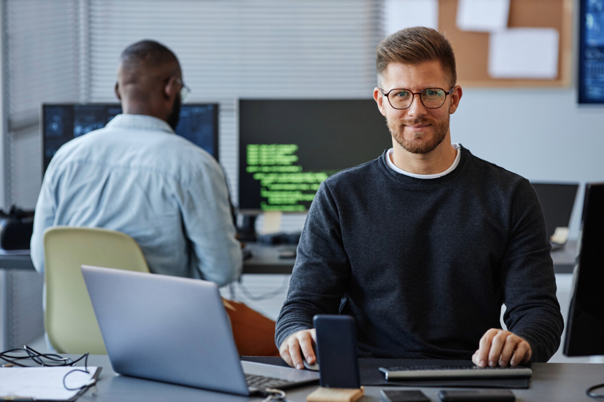 A man wearing glasses works at a desk with a laptop, keyboard, and smartphone, possibly overseeing his Shopify store. Meanwhile, another person is busy at a computer in the background, probably handling DTC ecommerce tasks.
