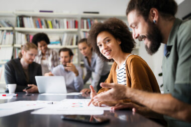 In an office setting, a group of people is gathered around a table filled with documents and electronic devices. Two individuals, honing in on their DTC strategy, engage in discussion, while others blur into the background.
