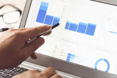 A person uses a pen to point at data charts on a laptop screen, revealing various blue bar graphs and pie charts, crucial for optimizing ecommerce strategies on platforms like Shopify.