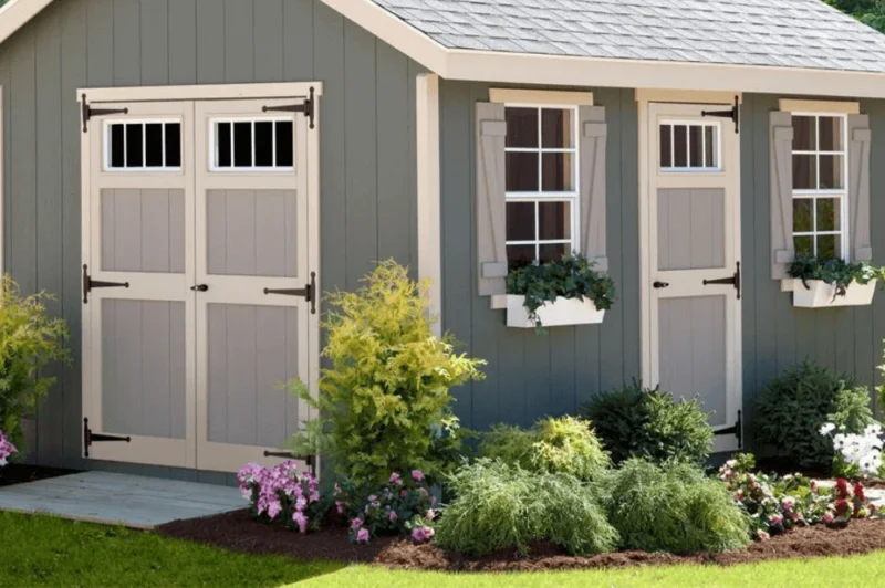 A garden shed with gray siding, beige trim, and double doors stands ready for your tools or latest DTC venture. Adorned with windows, shutters, and charming flower boxes, it gracefully complements the vibrant array of plants and flowers in the surrounding garden.