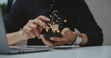 A person navigates their smartphone, where a digital shopping cart icon floats alongside symbols of communication, embodying seamless ecommerce. Nearby, a laptop rests on the table, ready to enhance this Shopify-driven experience.