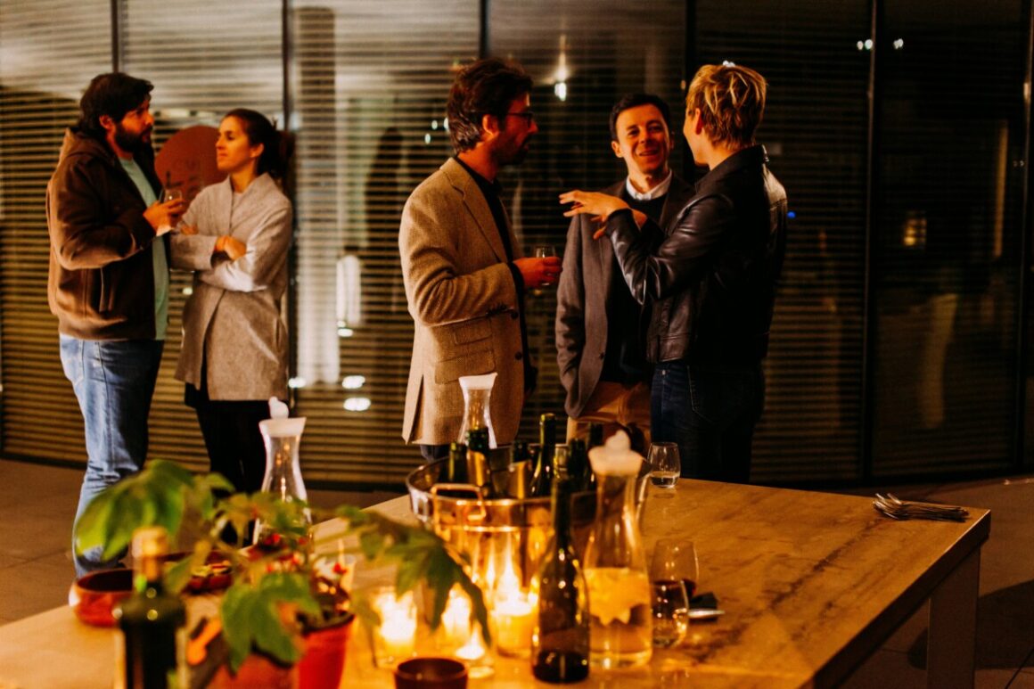 Five people socializing with drinks at a dimly lit gathering, standing near a table adorned with candles and bottles, reminiscent of a Shopify team celebrating their latest DTC success.