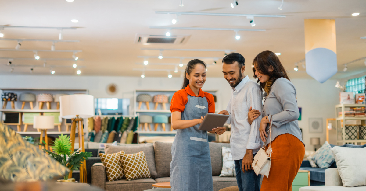 A store employee shows a tablet to a smiling couple in a furniture showroom, highlighting their selection of sofas and cushions available through their Shopify-powered ecommerce platform.