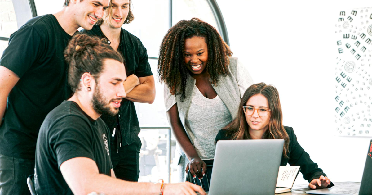 Five people gather around a laptop, engaged in a collaborative discussion about DTC strategies in a brightly lit room, exploring innovative ecommerce solutions.