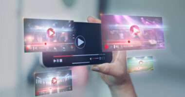 A hand holds a smartphone displaying a video player interface. Surrounding the phone are floating video thumbnails with play icons and progress bars, illustrating how DTC brands leverage ecommerce platforms to enhance their storytelling through dynamic visual content.