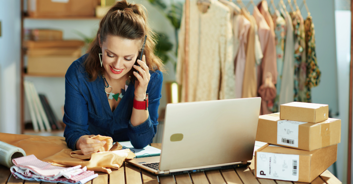 A woman in a blue blouse discusses small business options over the phone, glancing at fabric. Her laptop and parcels rest on the table, and clothing racks fill the background, setting the scene for her creative enterprise.