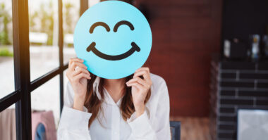 Person holding a blue smiley face sign in front of their face indoors, showcasing simple methods to boost customer satisfaction with cheerful charm.
