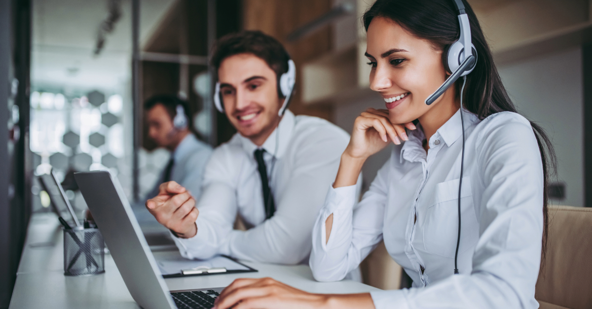 Two call center employees wearing headsets work at computers, one pointing at a screen related to Shopify activities, both smiling.
