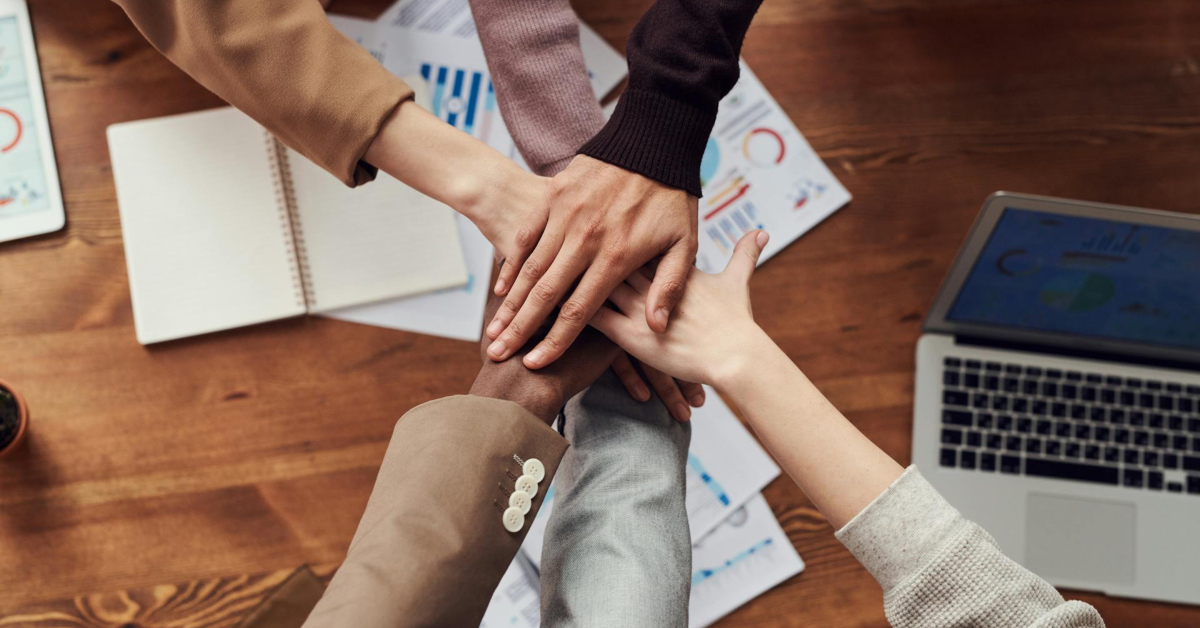 A group of hands placed together over a wooden table with laptops, documents, and a notebook in the background symbolizes ecommerce teamwork and collaboration, essential for successful Shopify DTC strategies.