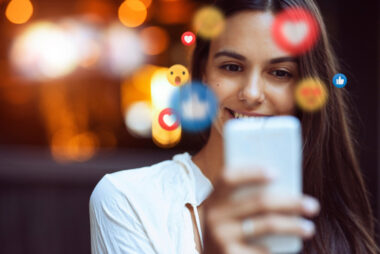 A person smiles while looking at a smartphone, with social media reaction icons like hearts and thumbs-ups floating around, reflecting their excitement about the latest DTC ecommerce trends. Blurred background.