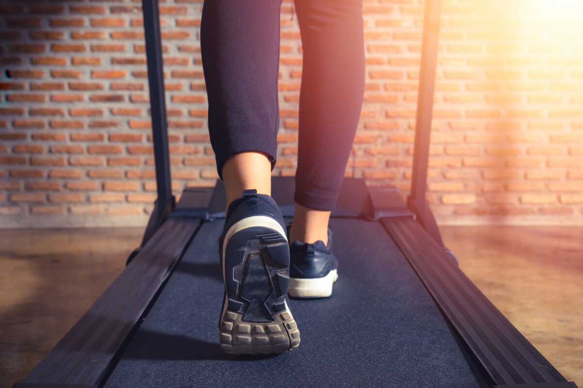 A person walks on a treadmill, seamlessly integrating sustainable fitness into their routine. Dressed in black athletic shoes and pants, they move with purpose against the backdrop of an industrial brick wall.
