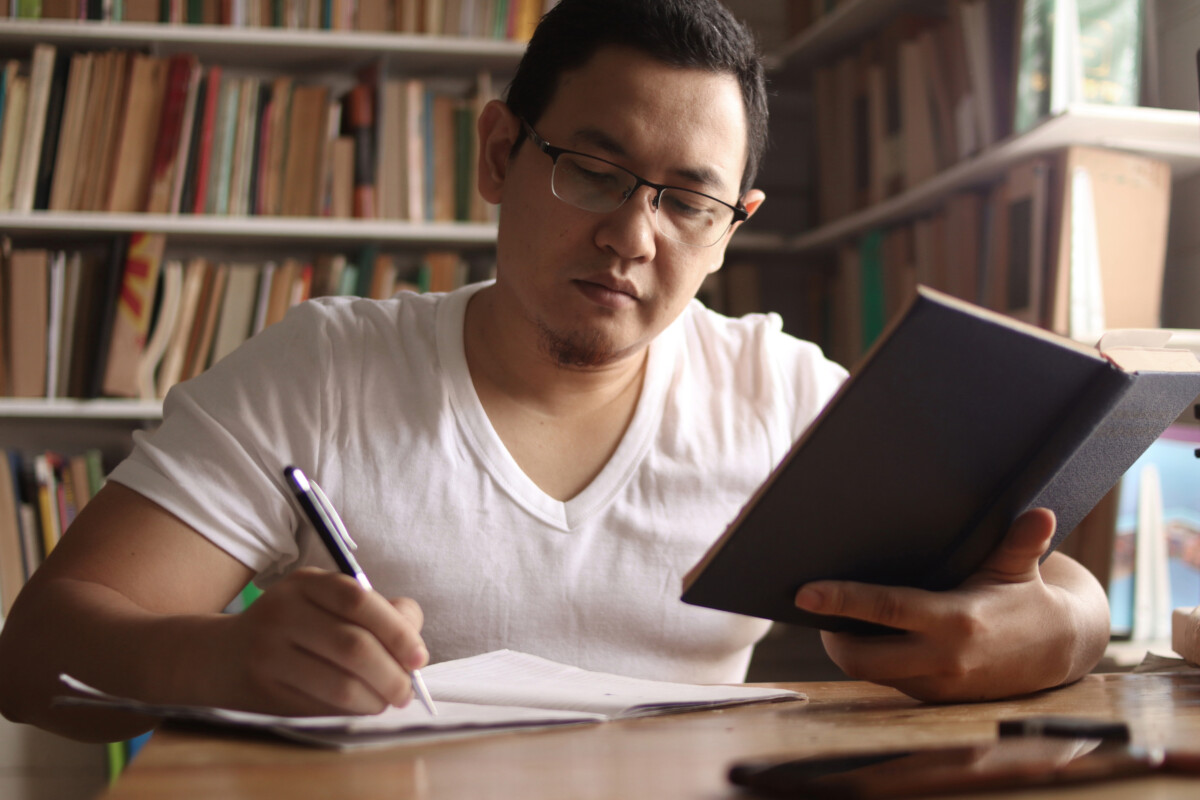 Seated at a wooden table, a person in a white shirt carefully writes in a notebook while holding a book, thoughtfully incorporating an exam preparation strategy. Bookshelves line the background, hinting at the depth of knowledge within reach.