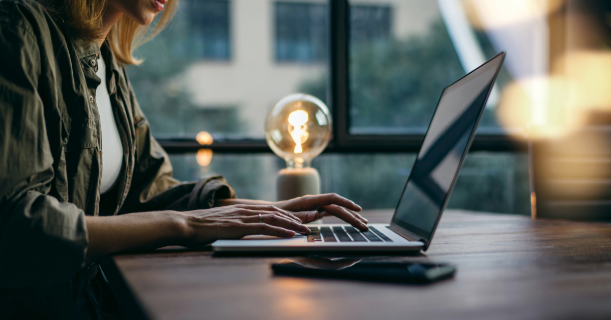 A person is typing on a laptop at a wooden desk, illuminated by a lit bulb lamp, planning the next big move in their ecommerce strategy. A smartphone rests nearby, ready to assist in their Shopify or DTC endeavors.
