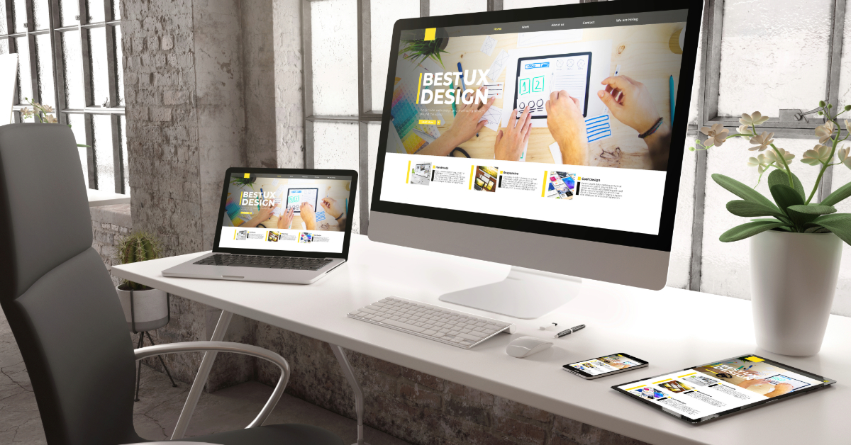 A modern workspace equipped with a large desktop monitor and laptop showcasing "Best UX Design" for ecommerce. A tablet and smartphone rest nearby, ready for DTC strategies. Plants and a window infuse the area with a bright atmosphere.