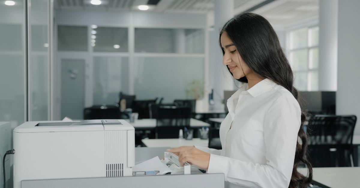 A woman in a white shirt uses a printer in an office buzzing with ecommerce energy. She is inserting paper into the machine, surrounded by desks and chairs where Shopify strategies and DTC dreams come to life.