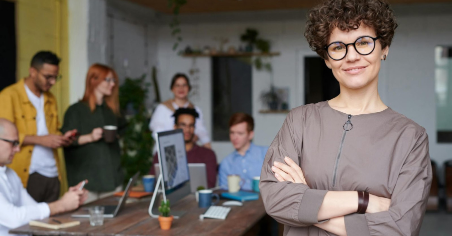 A smiling person with curly hair and glasses stands confidently, embodying the essence of DTC strategy, as a group collaboratively works at a table in a bright room.