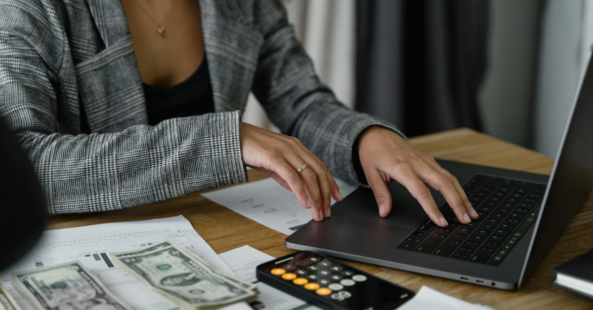 A person in a plaid blazer is using a laptop, surrounded by documents, cash, and a calculator on a wooden table, meticulously tracking monthly costs.