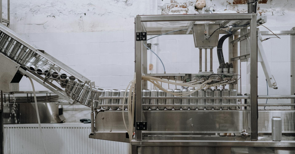 In a bustling factory, the industrial machine operated by the right manufacturer expertly fills metal cans with liquid. Surrounded by an intricate network of metal pipes, with white tile walls as the backdrop, this setup exemplifies precision and expertise in product manufacturing.