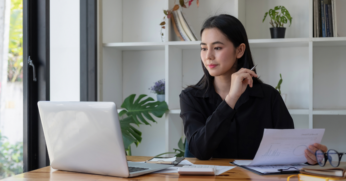 A woman in a black shirt sits at a table with a laptop, holding papers and a pen, looking thoughtful. She's likely researching the best planning consultants near me to ensure her project succeeds.